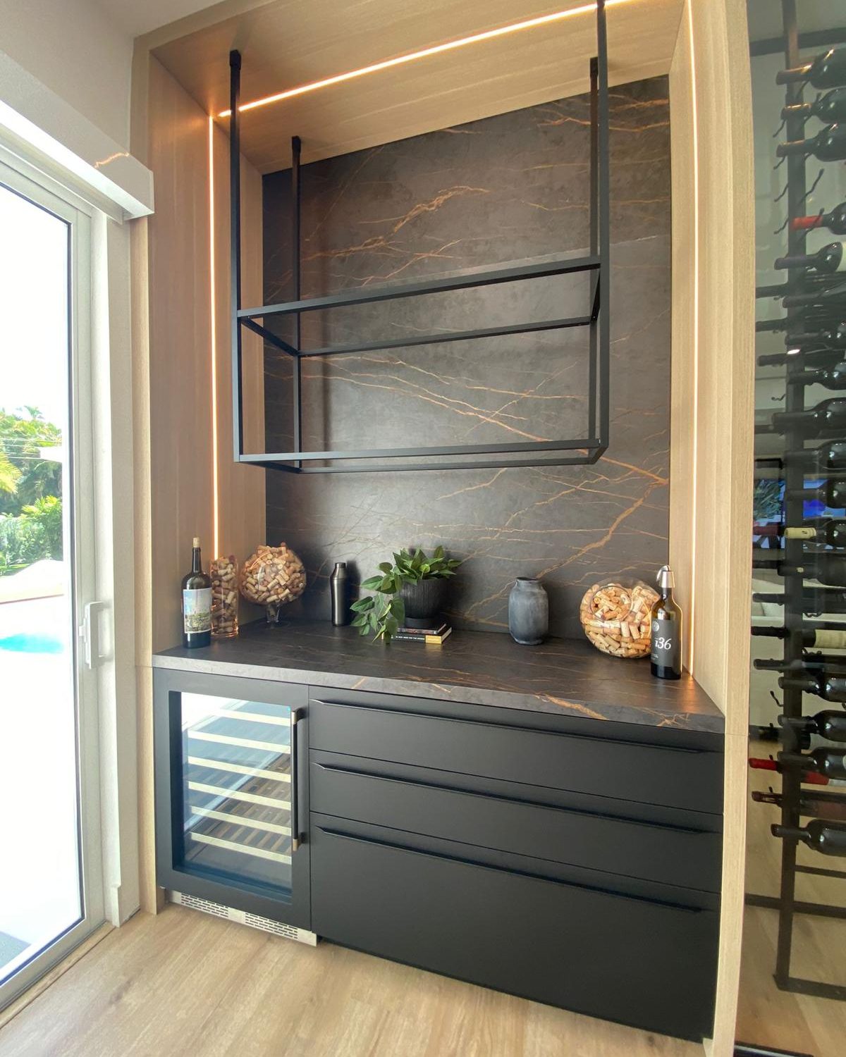 Atmospheric view of a custom dry bar with cove lighting, dark marble back wall, and glass-enclosed wine room beyond