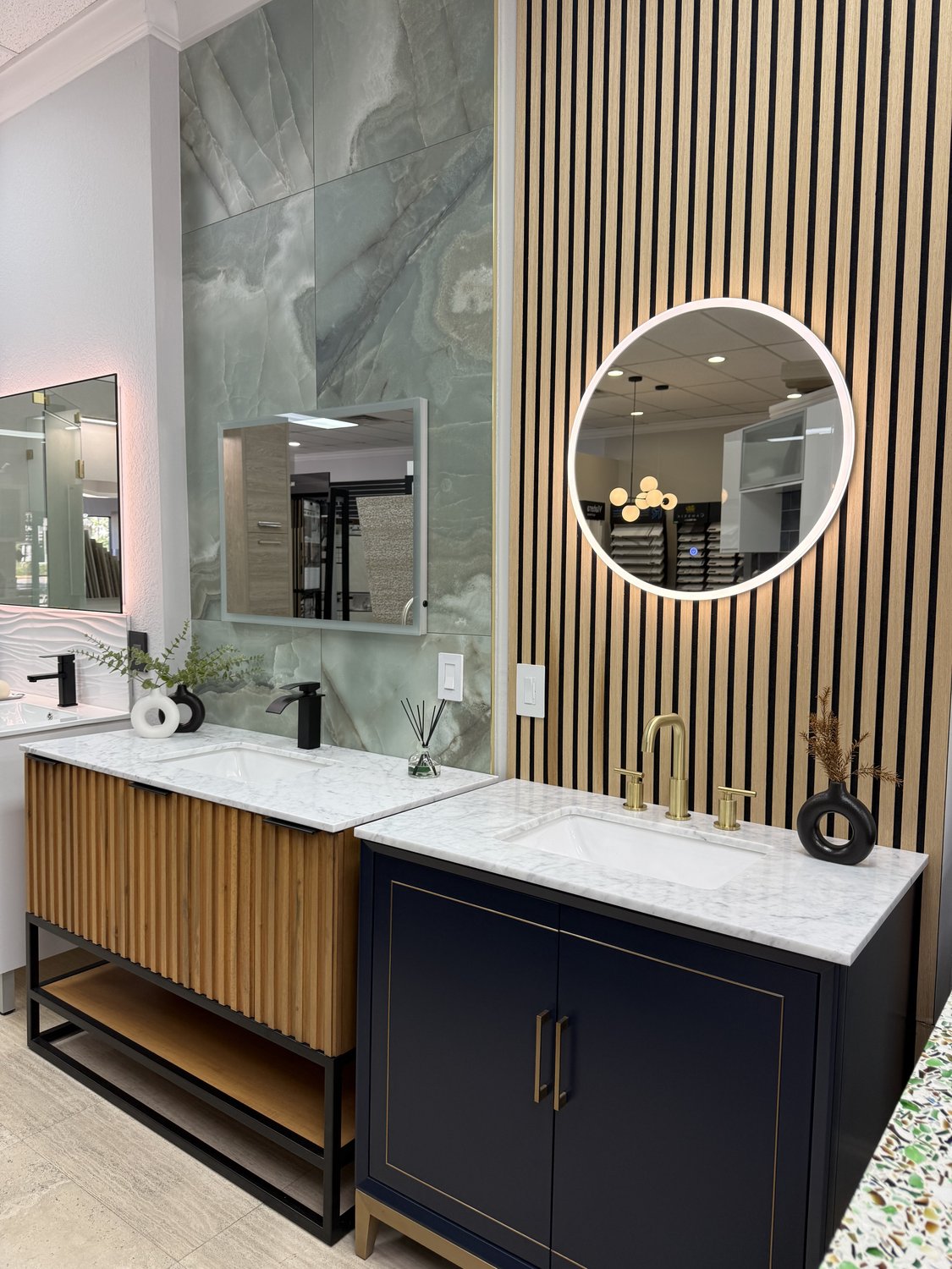 Two vanity vignettes inside the Acassa showroom: a fluted oak vanity beside a navy vanity with brass fixtures, framed by a slatted wood wall and green onyx tile