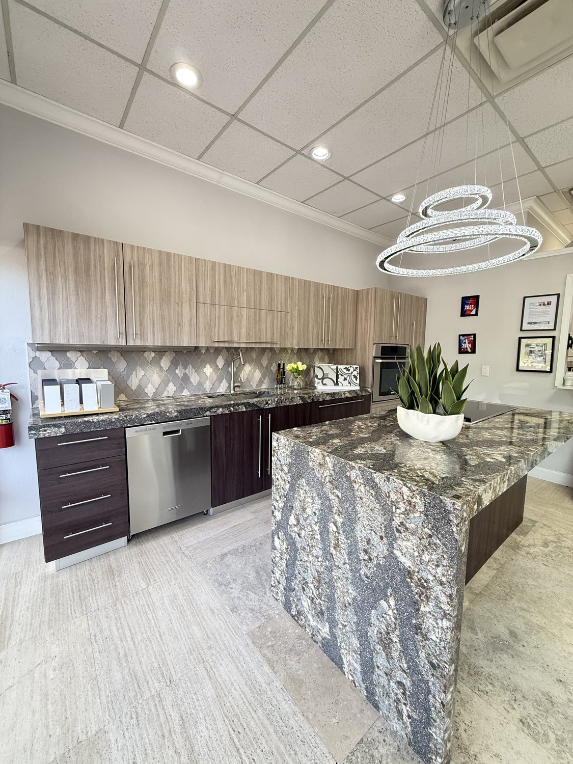 Showroom kitchen vignette with wood-grain cabinets, mosaic backsplash, and a heavily-veined stone waterfall island under a sculptural ring chandelier