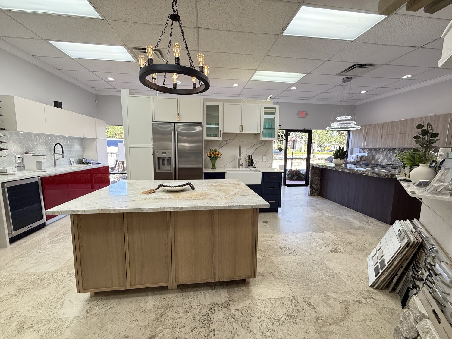 Wide view of the Acassa showroom, showing multiple full-scale kitchen vignettes, stone-slab samples, and travertine flooring under a forged-iron chandelier