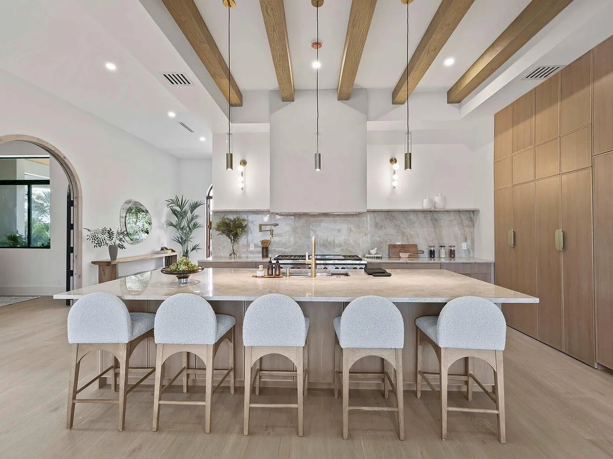 Open kitchen with marble waterfall island, white boucle bar stools, exposed wood ceiling beams, and arched doorway to the dining room
