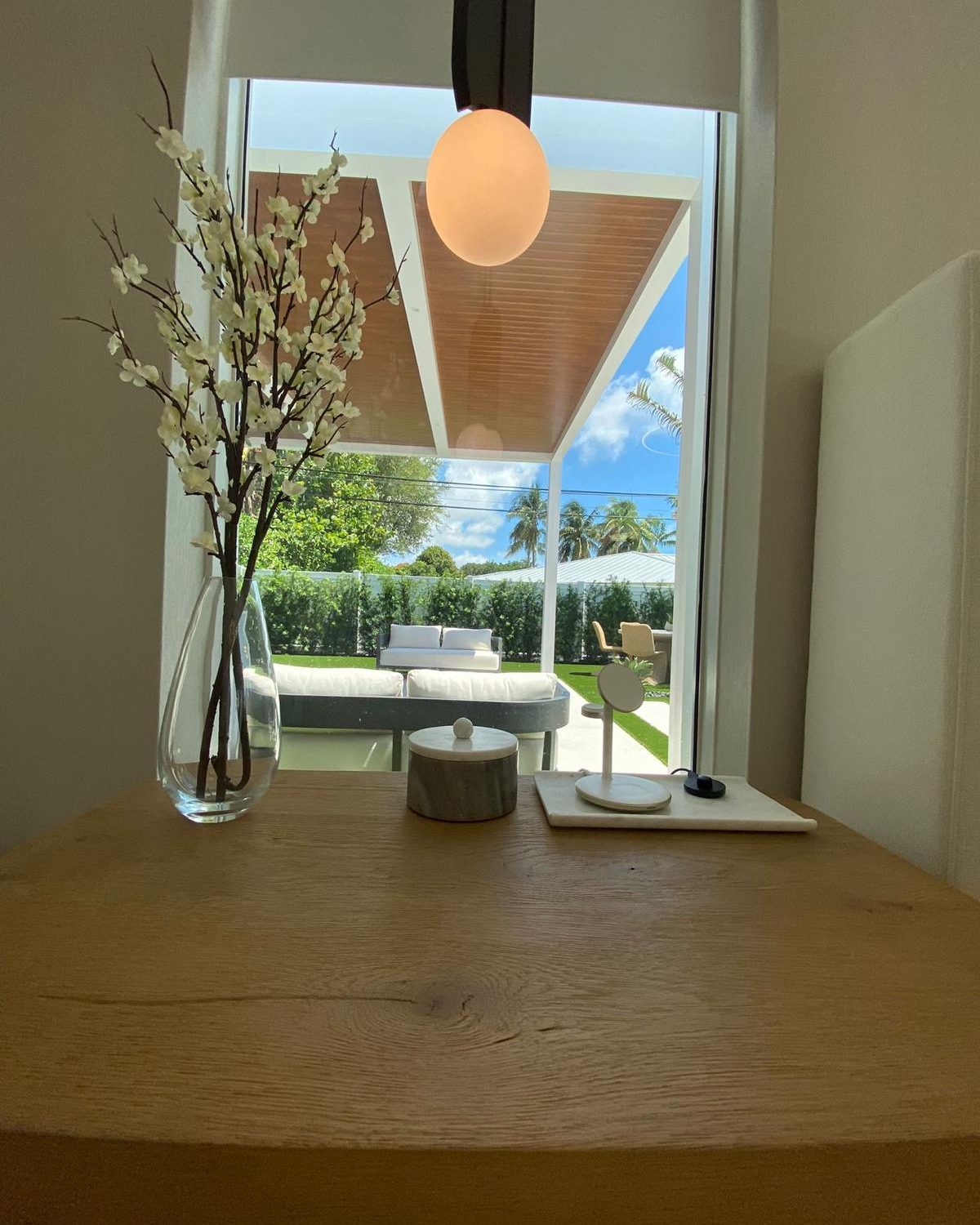 View from inside the home through window to backyard pergola, lounge furniture and palm trees